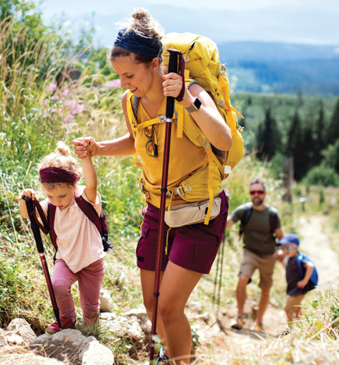 Lady, holding hand of small child, hiking up a hill.  Both are using hiking poles to navigate hill.  