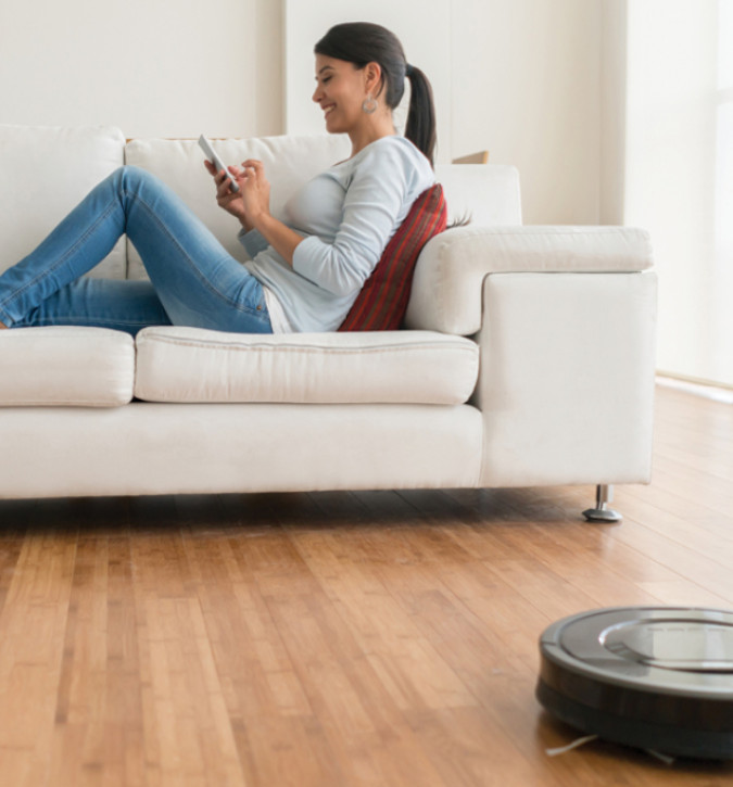 Lady sitting on couch, looking at her phone and smiling while rumba vacuum cleans floor
