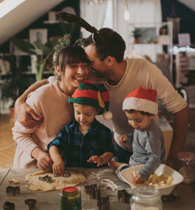Man and lady standing behind two small children. All standing at counter rolling cookie dough and using cookie cutters. Smiling and wearing Christmas hats.