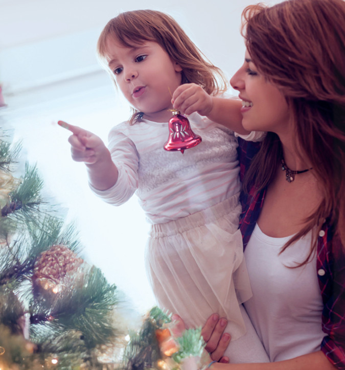 Smiling female with long hair holding little girl who is all dressed up.  Little girl is holding a bell and pointing at Christmas tree